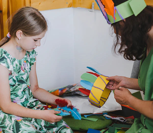Девочка с преподавателем делают игрушечную корону из перьев и бумаги.