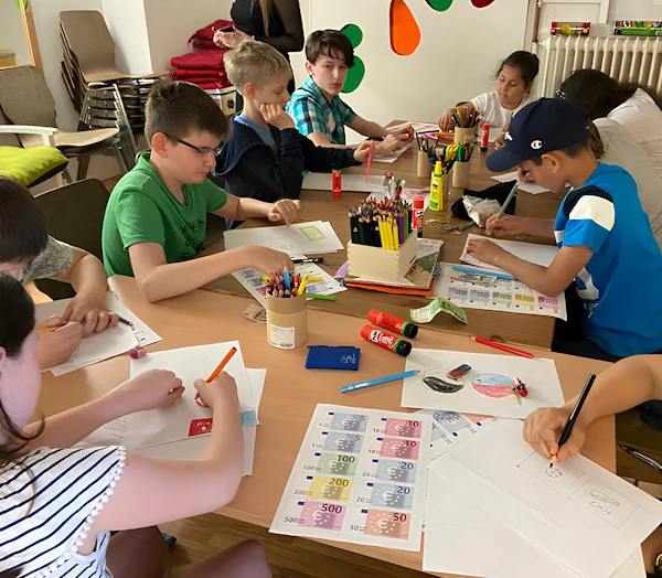 Группа детей сидит за столом и рисует придуманные ими банкноты.