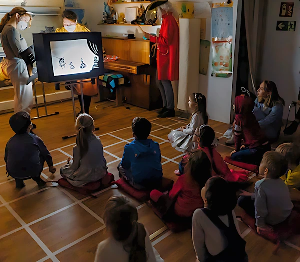 Две преподавательницы показывают группе детей младшего возраста театр теней. Дети сидят на полу и с восторгом смотрят представление на экране.