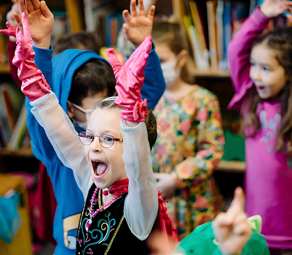 Дети в нарядных костюмах веселятся на мероприятии в Чемодане сказок. Они радуются и смеются, кружатся с поднятыми вверх руками. На переднем плане девочка в ярких розовых перчатках.